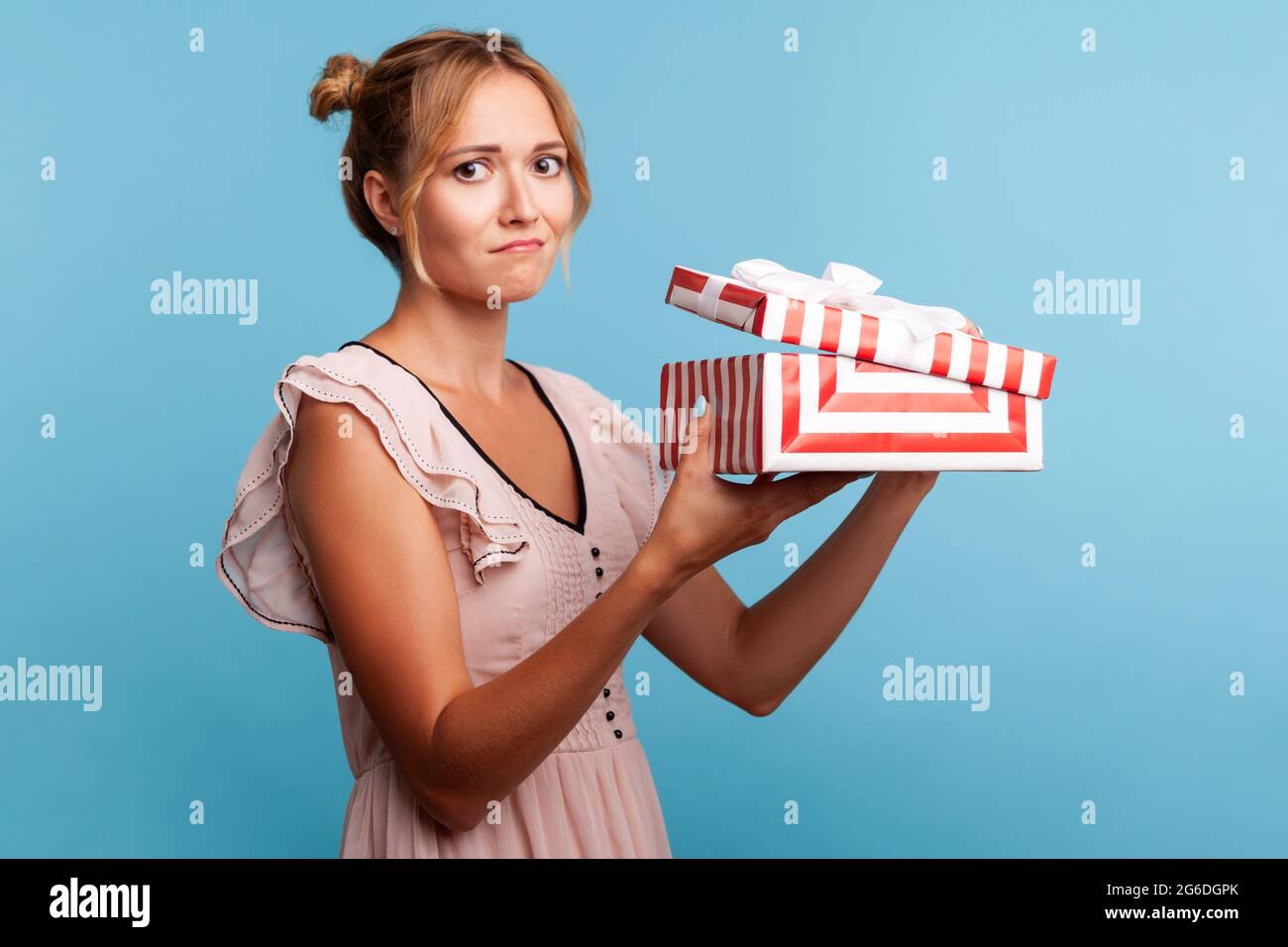 Portrait of upset young adult blonde female with bundles, holding red striped present box, has sad expression, being disappointed with birthday gift. Stock Photo