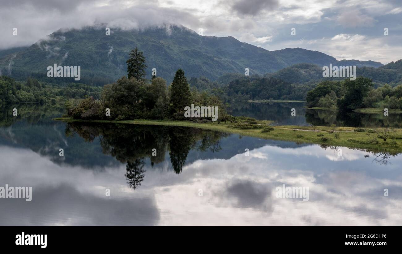 Loch Achray, Loch Lomond and Trossachs National Park, Scotland, UK. 4 July 2021. Pictured: Loch Achray turns into a mirror reflection the areas outstanding natural beauty with the Loch Achray hotel in the background at the foot of the mountains.  Credit: Colin Fisher/Alamy Live News Stock Photo