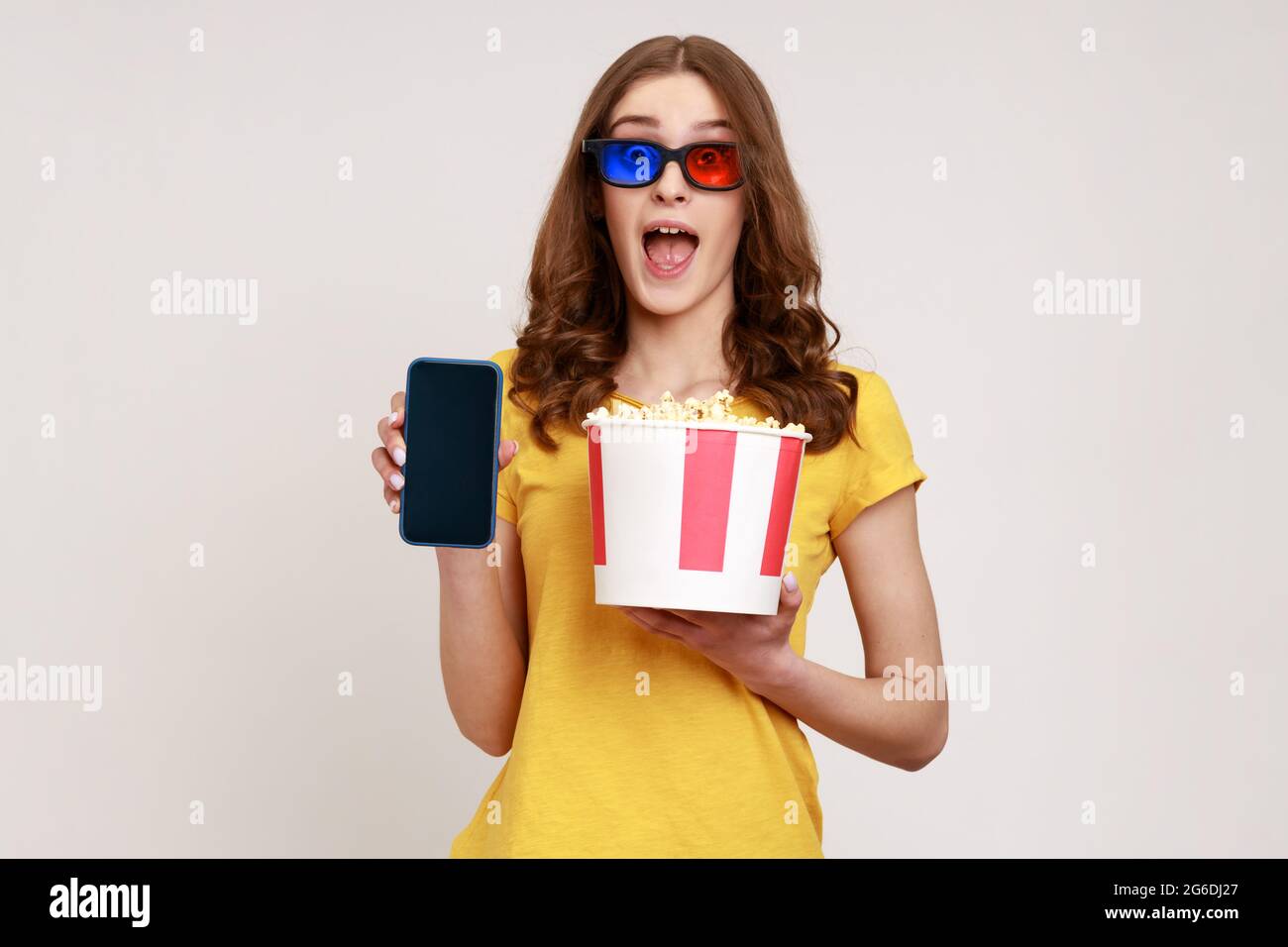 Excited young beautiful woman in yellow casual style t-shirt and 3d glasses holding bucket popcorn and mobile phone with blank display. Indoor studio Stock Photo