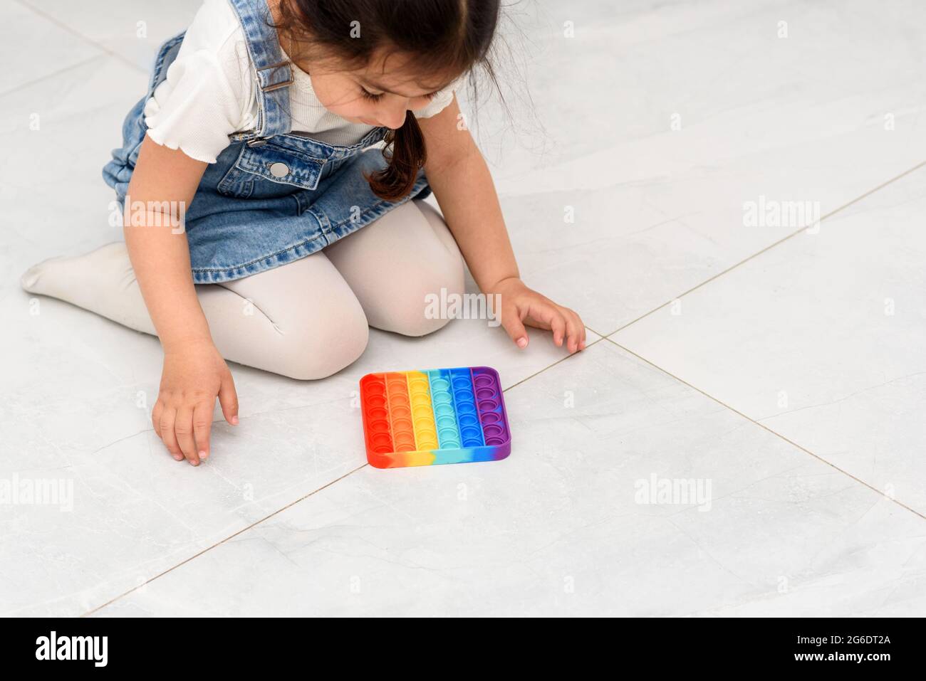 Young Little Girl Sitting On The Floor At School Playing Poppit-New Fidget Toy, Popular With Kids, Helps Them To Concentrate. Child Playing With The Pop It Fidget Toy. Stock Photo