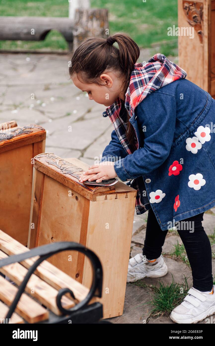 child girl helps parents polish an old wooden chest of drawers for repair and reuse against the background of a green garden, family hobby Stock Photo