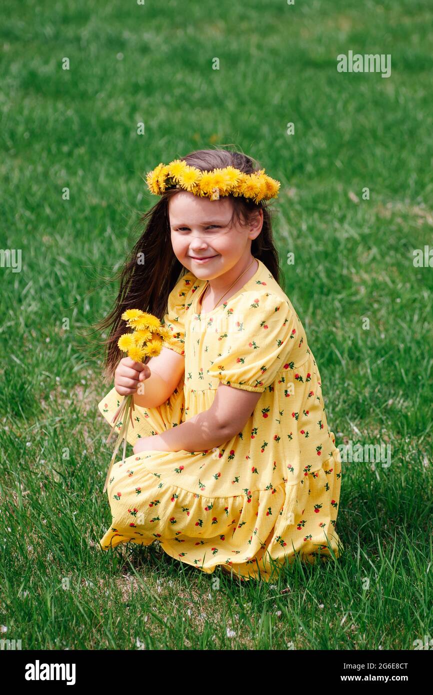 a full-length portrait of a beautiful child in a yellow dress sitting on the green grass and sniffing a bouquet of yellow dandelions Stock Photo