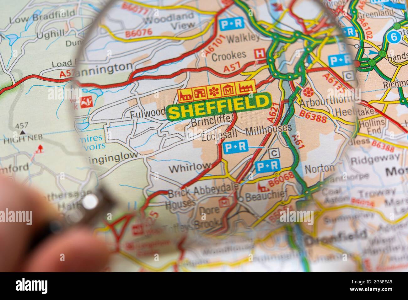 A macro closeup of a page in a printed road map atlas with a man's hand holding a magnifying glass showing an enlargement of Sheffield, England Stock Photo