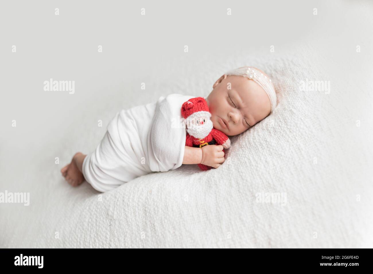 Newborn baby doll wrapped in a white wrap wearing a white pearled headband and cuddling a christmas santa claus lying on her side on a white blanket. Stock Photo