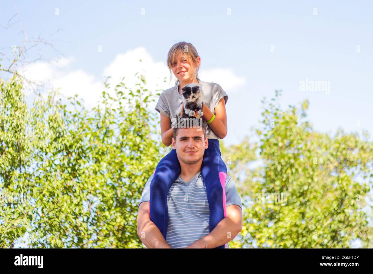 Defocused brother riding sister on back. Portrait of happy girl on man shoulders, piggyback. Girl holding cat, kitten. Family playing outdoor. Pet, an Stock Photo