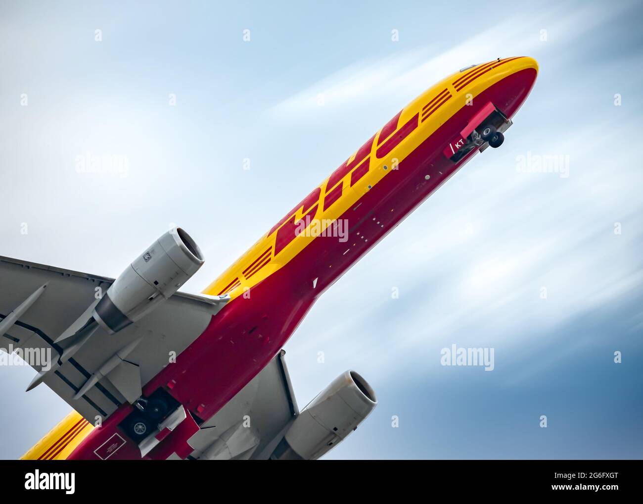 Yellow and Red DHL cargo aircraft jumbo jet taking off towards a blue sky with clouds blurring from motion. Wheels down after just lifting off runway. Stock Photo