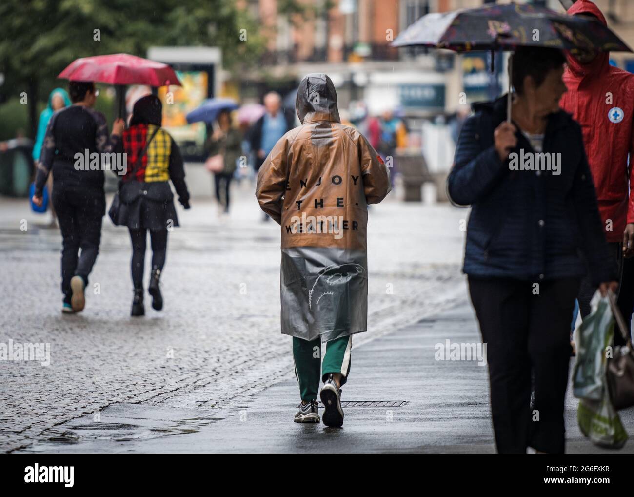 Enjoy the weather coat worn by high street shopper walking in depressing dark wet gloomy town centre.  Umbrellas and typical English summer weather Stock Photo