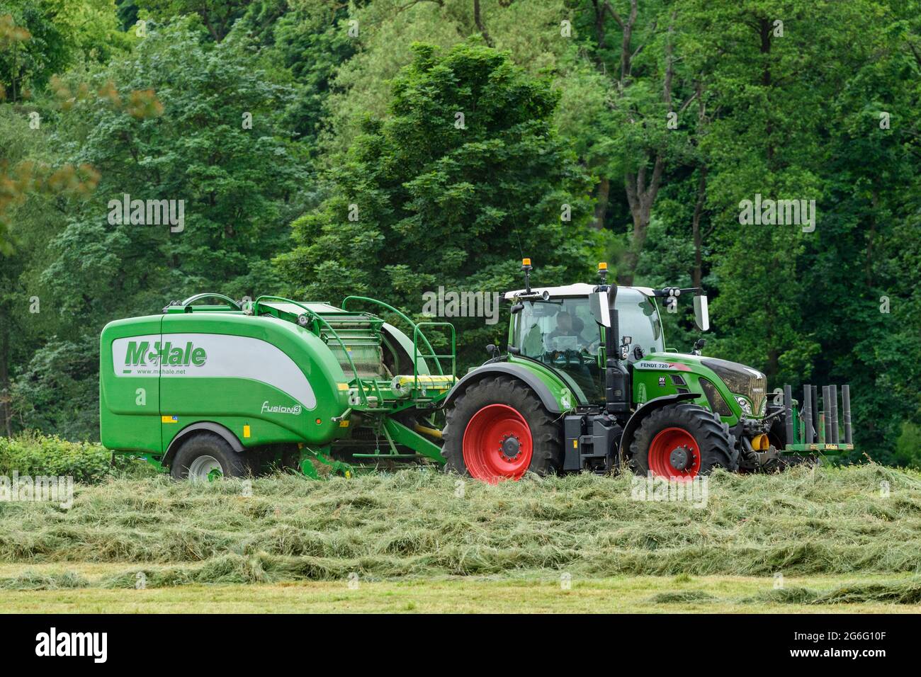 Hay or silage making (farmer driving green farm tractor at work in field, pulling McHale baler, collecting & baling dry grass) - Yorkshire England UK. Stock Photo
