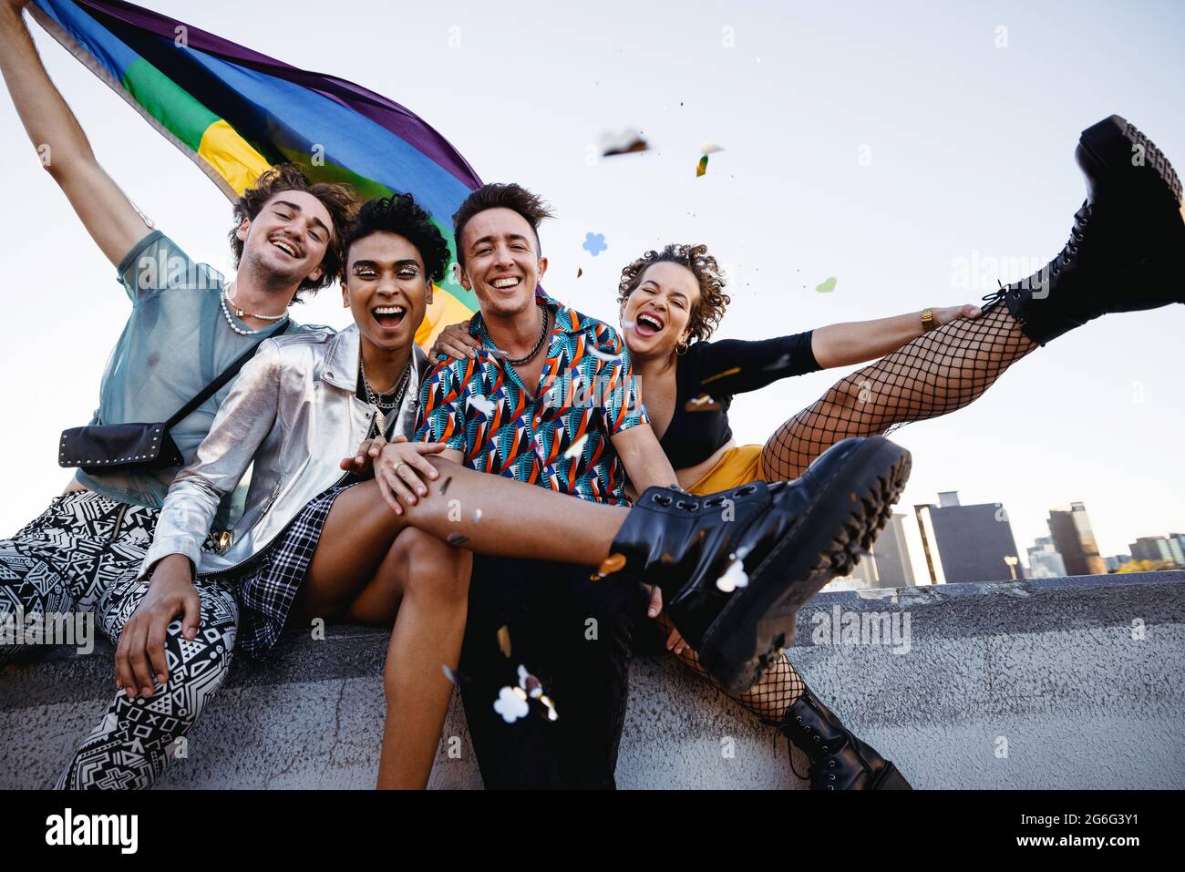 Friends celebrating gay pride while sitting together. Four members of the LGBTQ+ community smiling cheerfully while raising the pride flag. Group of q Stock Photo