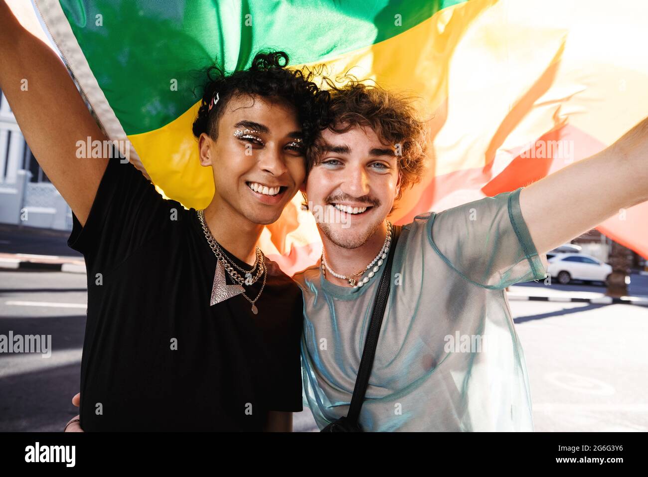 Two young queer men celebrating gay pride. Two young gay men smiling cheerfully while raising a rainbow flag at a gay pride parade. Two members of the Stock Photo