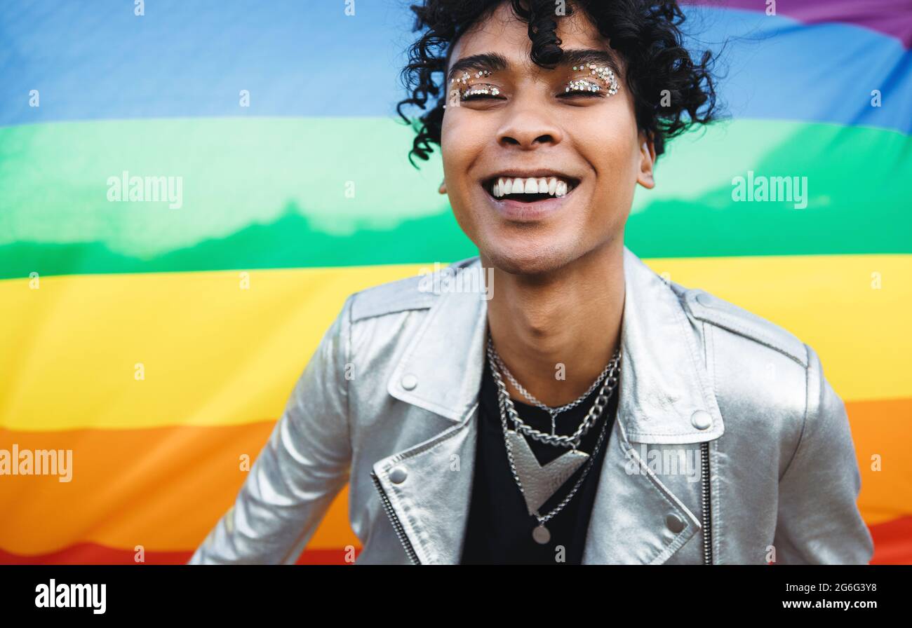 Young gay man embracing his queer identity. Happy gay man smiling cheerfully while standing against a pride flag. Gender non-conforming young man wear Stock Photo