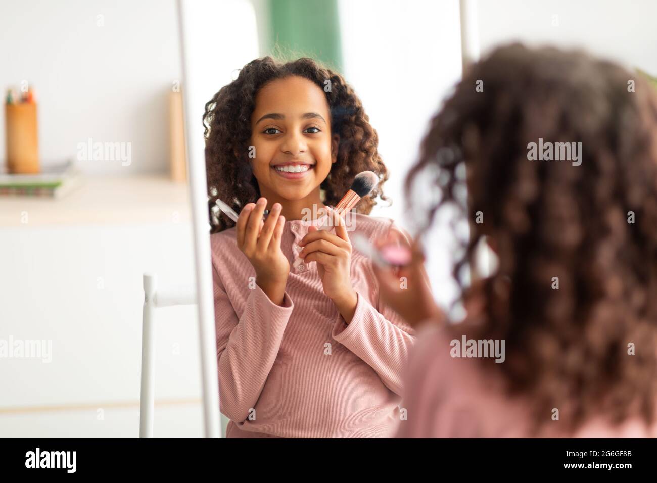 Home beauty salon concept. Happy african american girl doing makeup, holding brushes and smiling at mirror Stock Photo