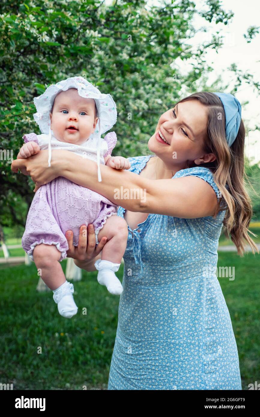 Young woman with loose fair hair rocks in arms baby girl and plays with kid  standing in park against green trees in white blossom Stock Photo - Alamy