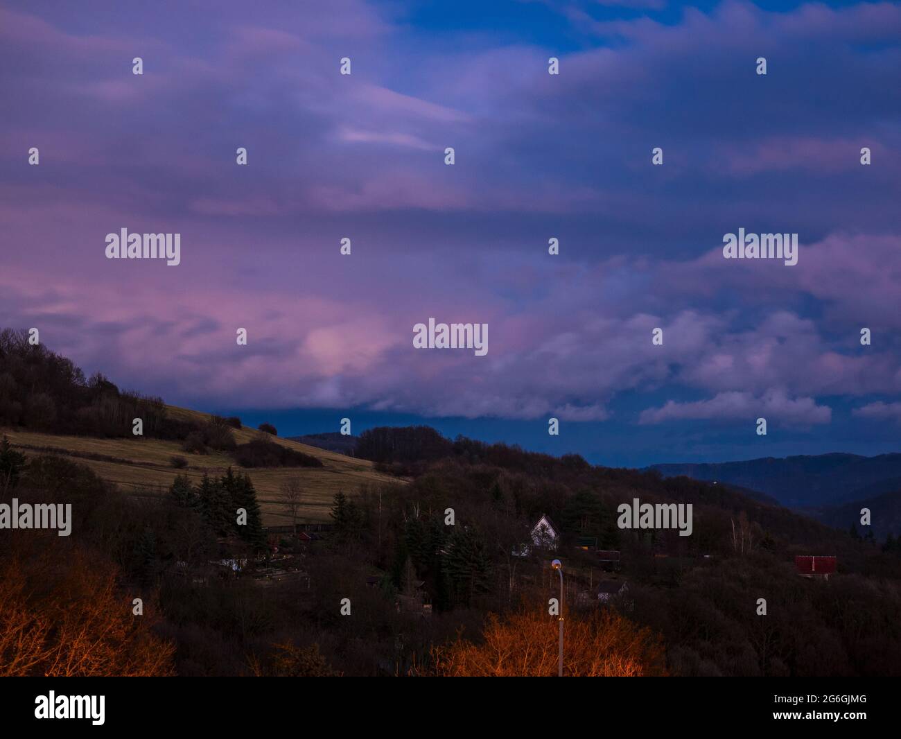 Massive clouds over hilly landscape in winter without snow, with shining street lamp in the foreground and tiny lights in the garden in the background Stock Photo