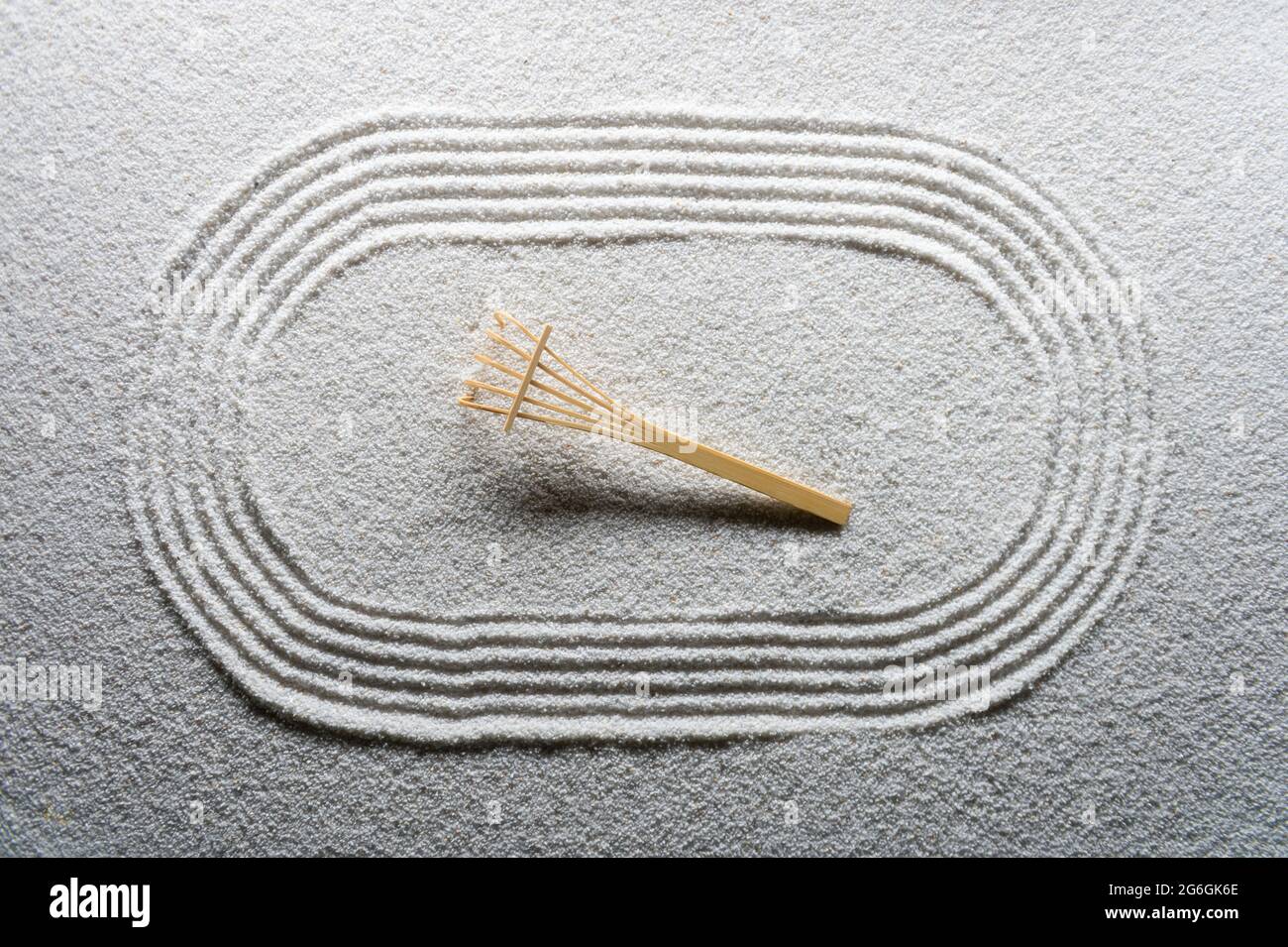 Small wooden rake sits on Japanese Zen garden raked with a simple sports running track oval in textured white sand Stock Photo