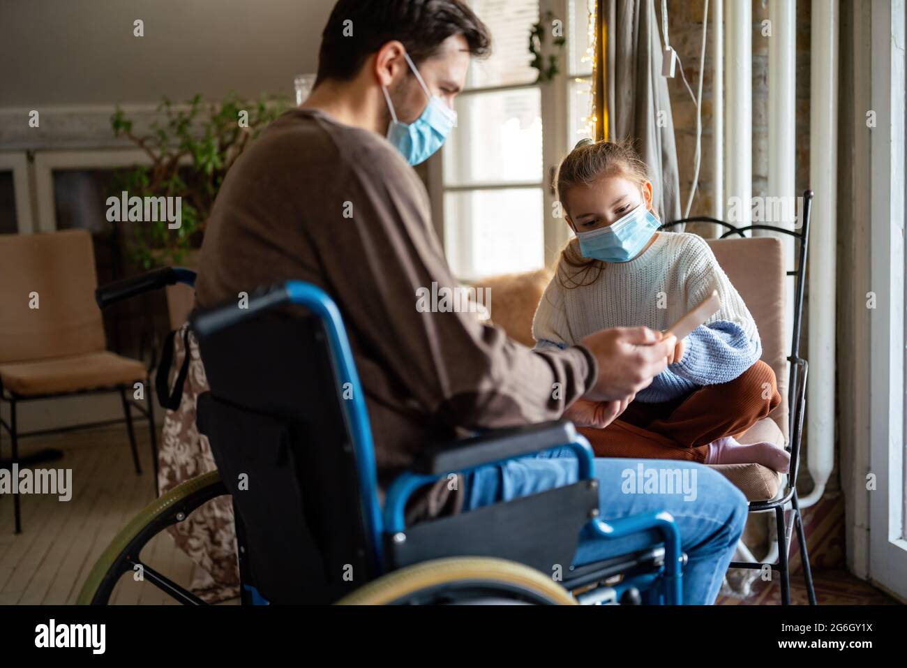 Father with disability in wheelchair using tablet at home with child while wearing masks. Stock Photo