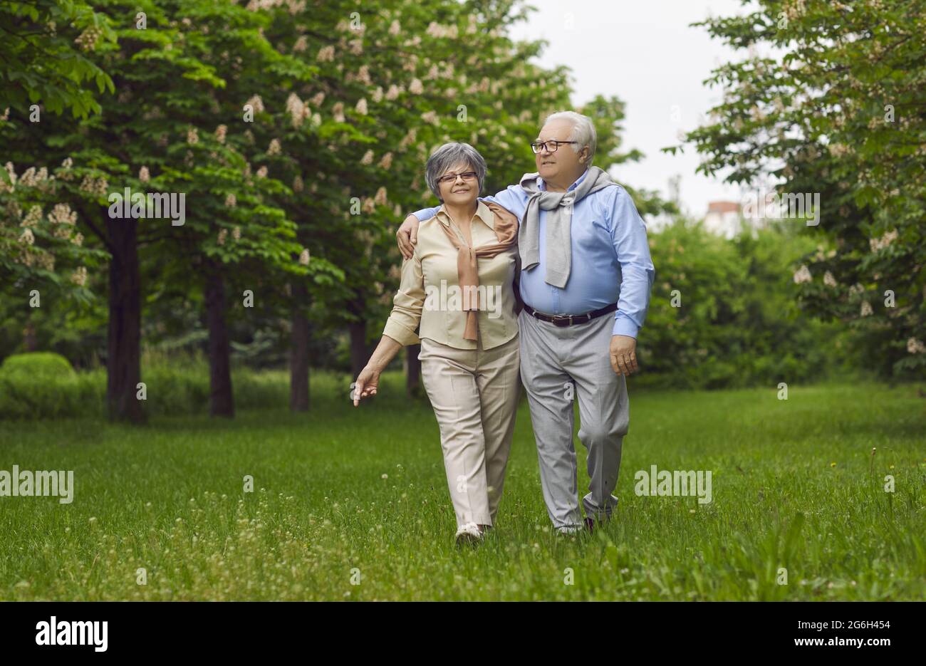 Senior man and woman are walking on green grass in the park among the trees. Stock Photo