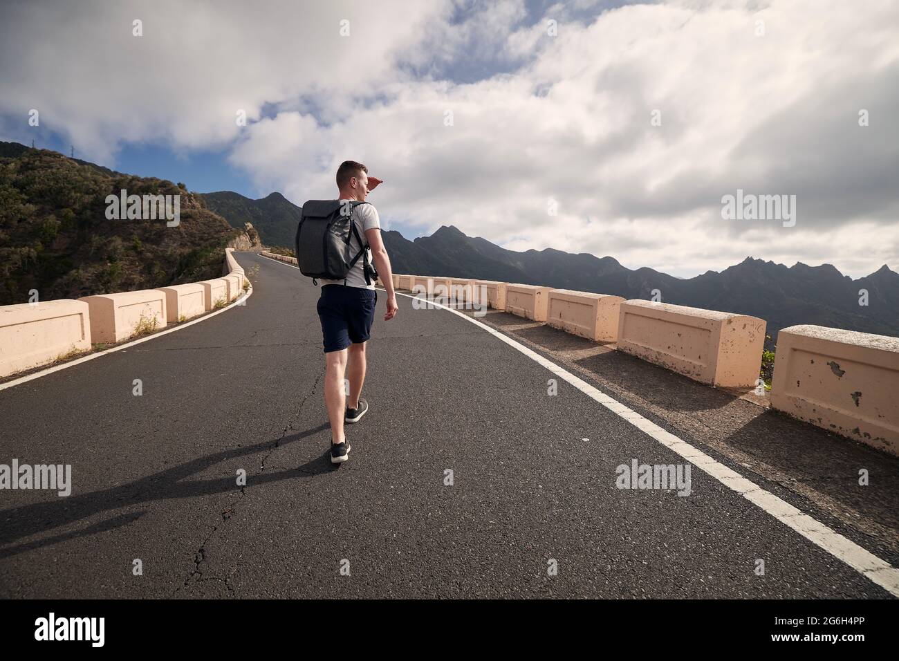 Rear view of man with backpack on mountain road. Young tourist enjoys trip on sunny day. Tenerife, Canary Islands, Spain. Stock Photo