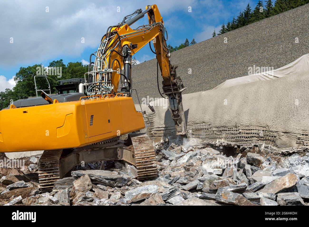 Excavator with hydraulic hammer chisels rock. Highway construction site on slope. The embankment is being secured. Wall with concrete and reinforcing Stock Photo