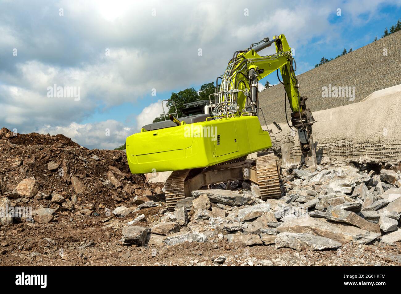 A yellow excavator with hydraulic hammer chisels rocks. Highway construction site on a slope. The embankment is being secured. Wall with concrete and Stock Photo