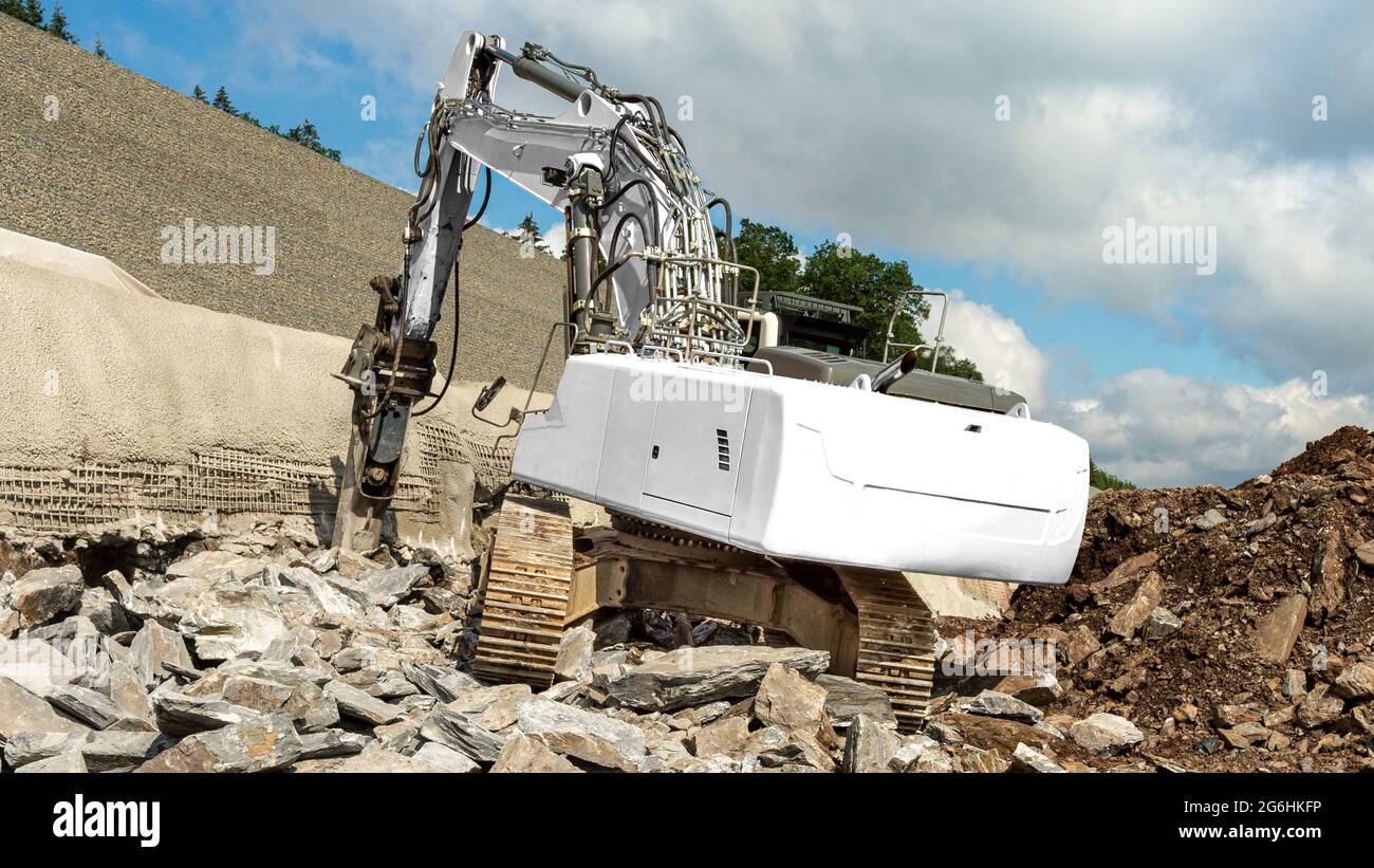 A white excavator offers space for own text. He chisels rocks with a hydraulic hammer. Highway construction site on slope. Wall with concrete and reba Stock Photo