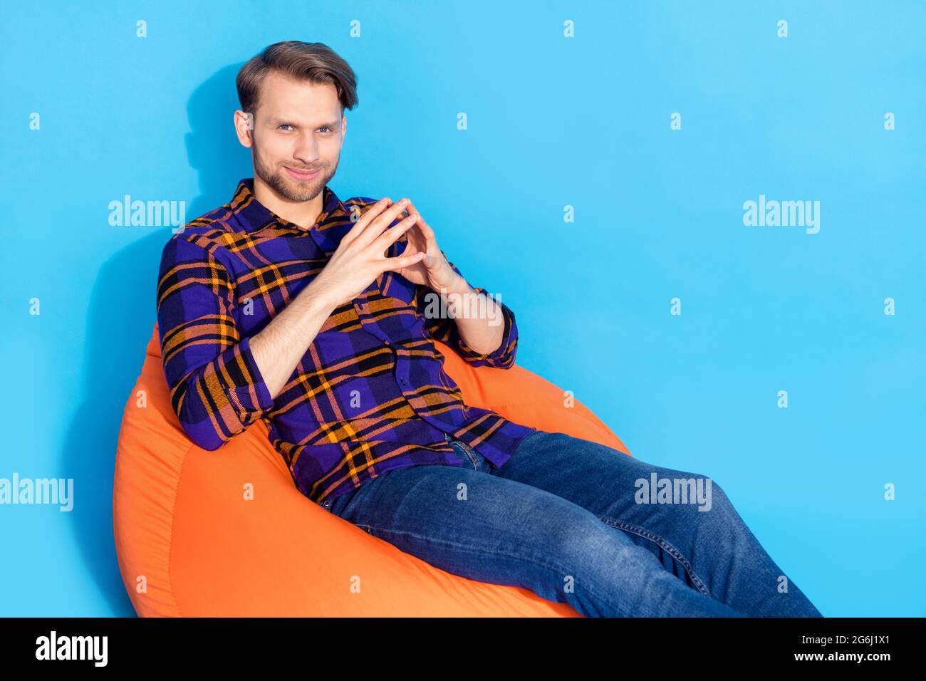 Photo of minded happy young man hold hands together evil genius sit bean bag isolated on blue color background Stock Photo