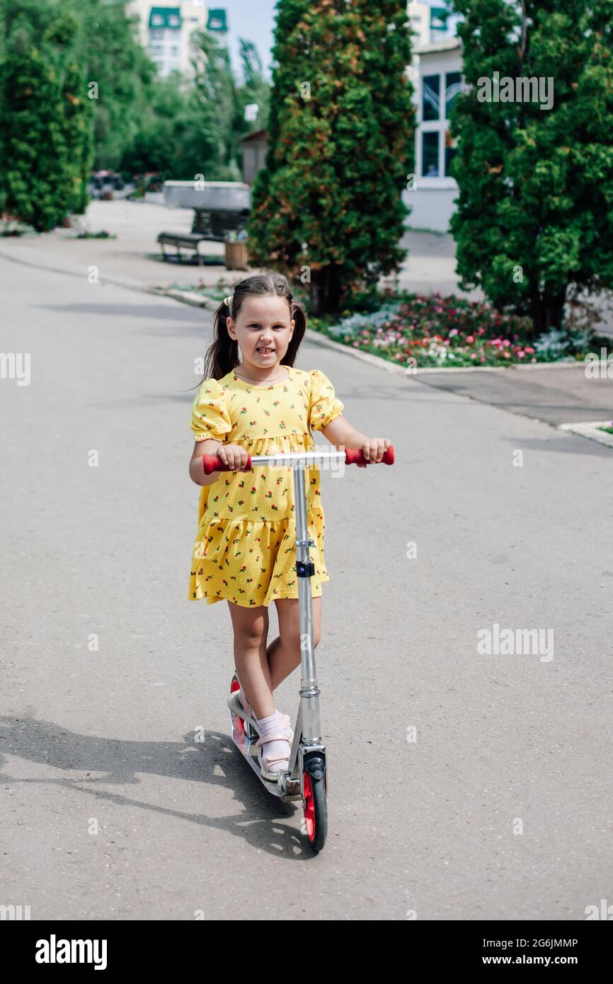 full-length portrait of a charming girl in a yellow summer dress riding a scooter on an asphalt path along an alley with cypresses Stock Photo