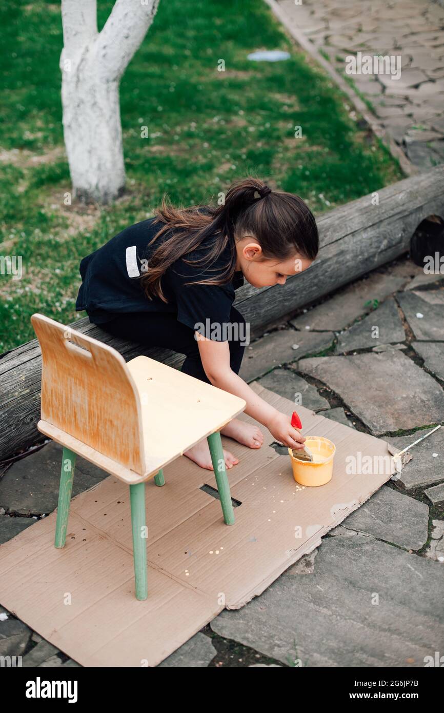 top view of a five-year-old girl dipping a brush in paint and about to paint a chair in a new color on a bench in the yard Stock Photo