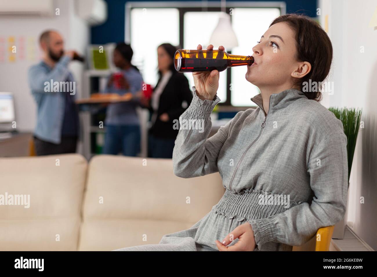 Cheerful woman drinking from bottle of beer after work at office with