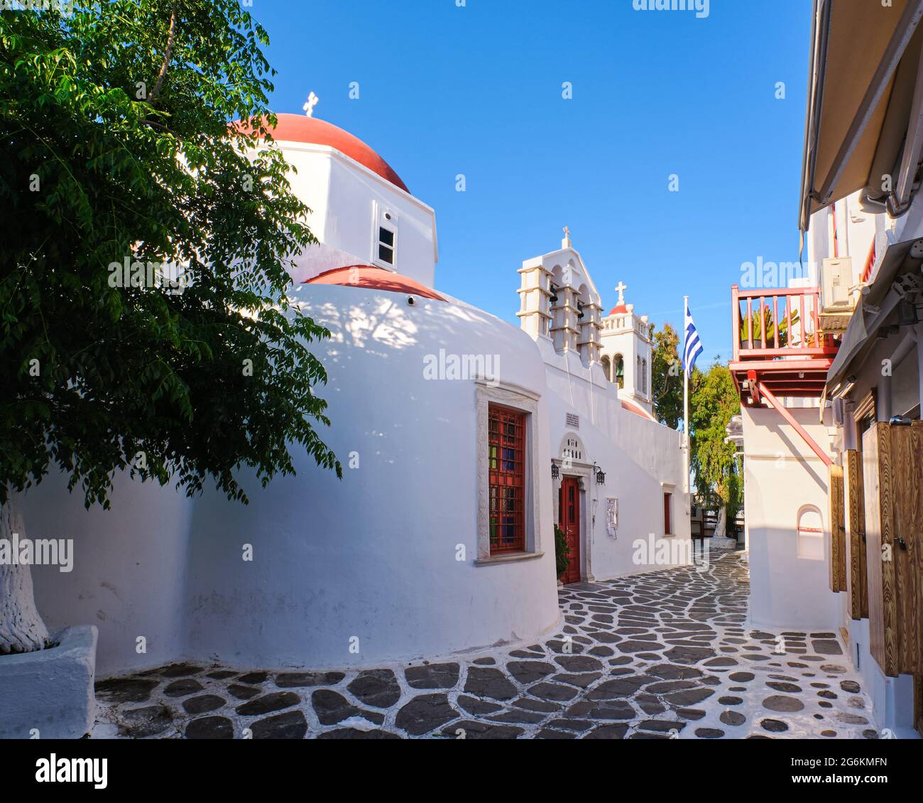 Traditional Greek Orthodox church in Greek island town. Red dome, whitewashed walls, Greek flag and bell tower. Mykonos, Cyclades, Greece. Stock Photo