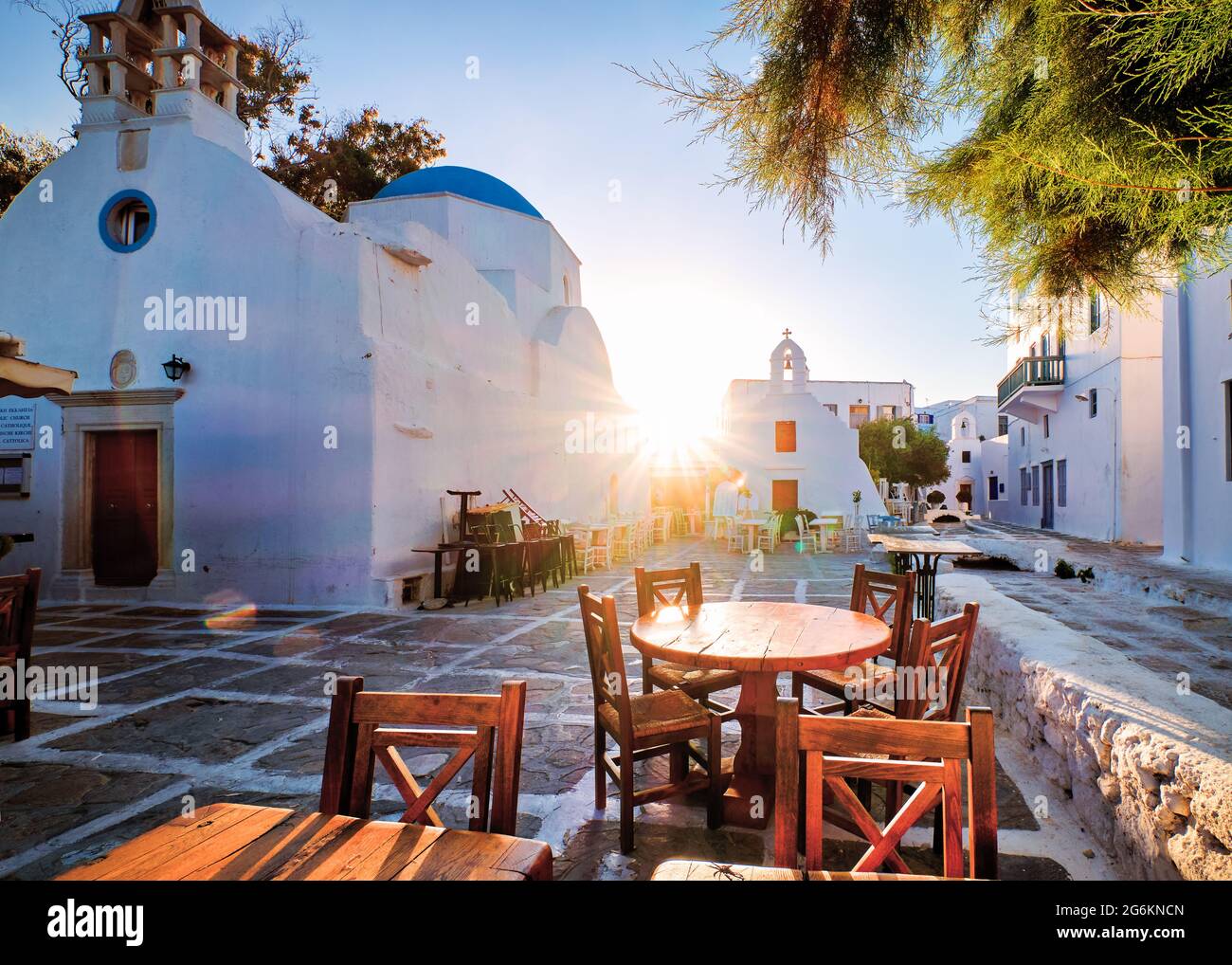 Sunrise in alleyways of Mykonos, Greece. Greek whitewashed churches in small town square, street cafe tables and chairs, sun and lens flares.  Stock Photo