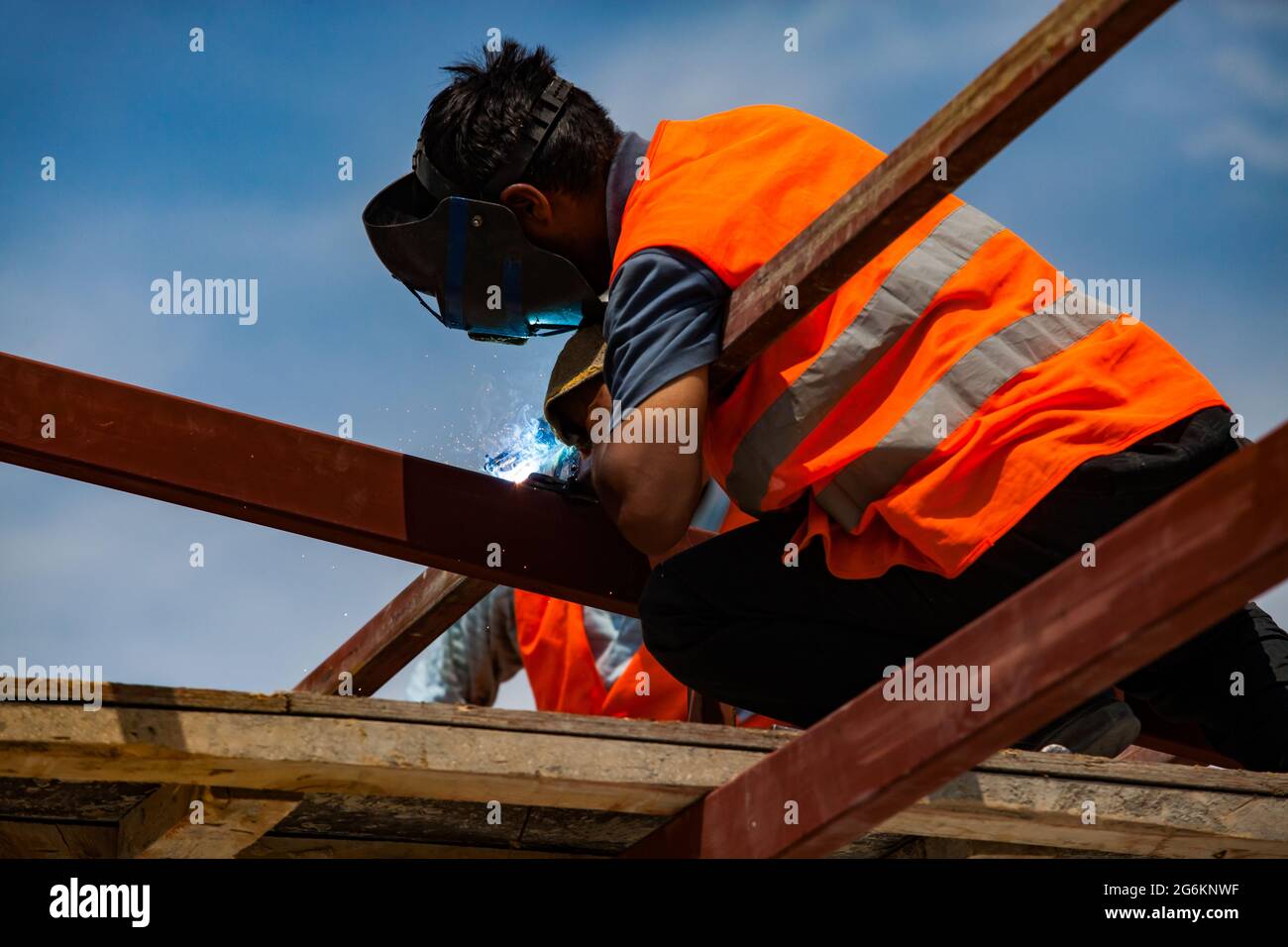 Close up photo of welder in welding mask and orange safety vest. No face. Flame in focus. Other part of photo blurred. Blue sky. Stock Photo