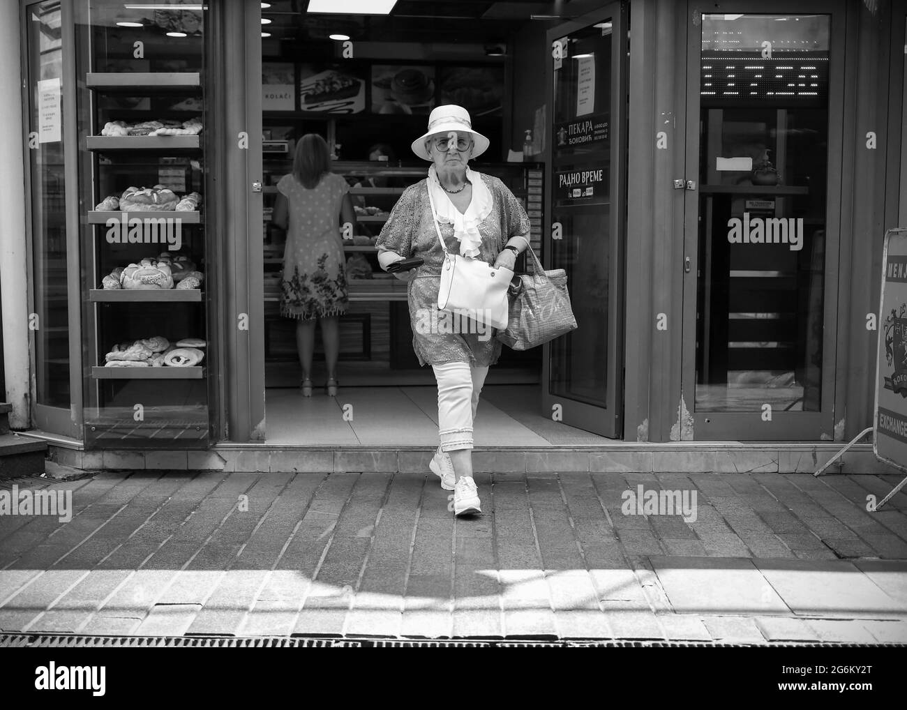 Belgrade, Serbia, Jul 3, 2021: An elderly woman wearing a white hat exits the bakery (B/W) Stock Photo