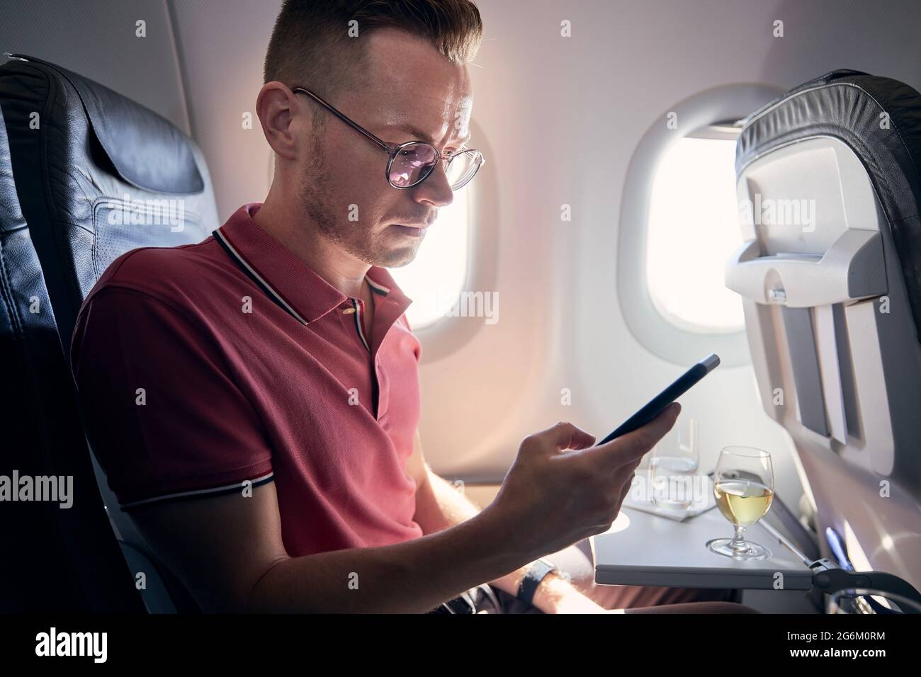 Young man using mobile phone at airplane. Passenger enjoys flight with internet connection a beverages. Stock Photo