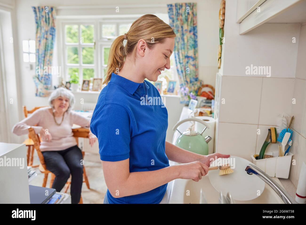 Female Home Help Cleaning House Doing Washing Up In Kitchen Whilst Chatting With Senior Woman Stock Photo