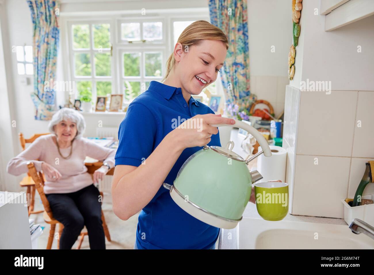 Female Home Help Making Cup Of Tea In Kitchen Whilst Chatting With Senior Woman Stock Photo