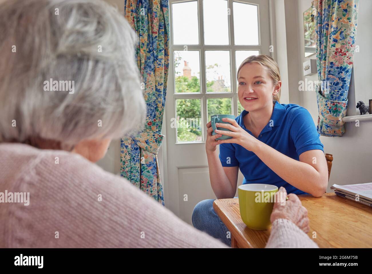 Female Home Help Having Cup Of Tea With Lonely Senior Woman In Kitchen Stock Photo