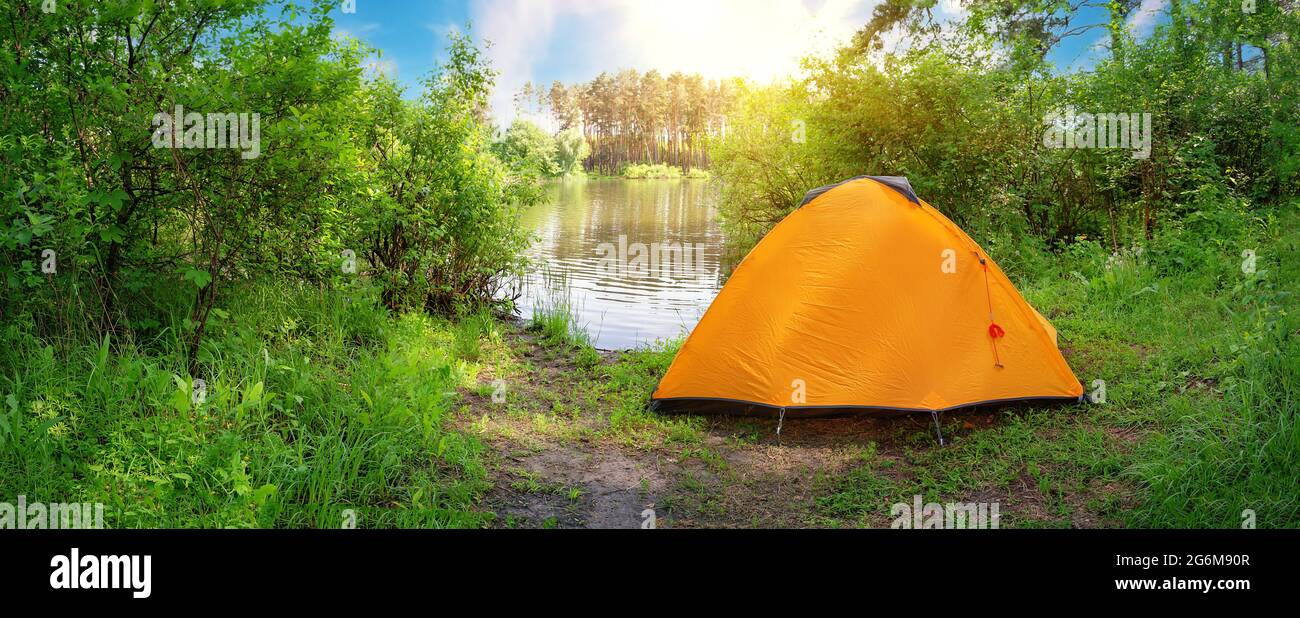 Orange tourist tent on the river bank in green foliage. Sunny day Stock Photo