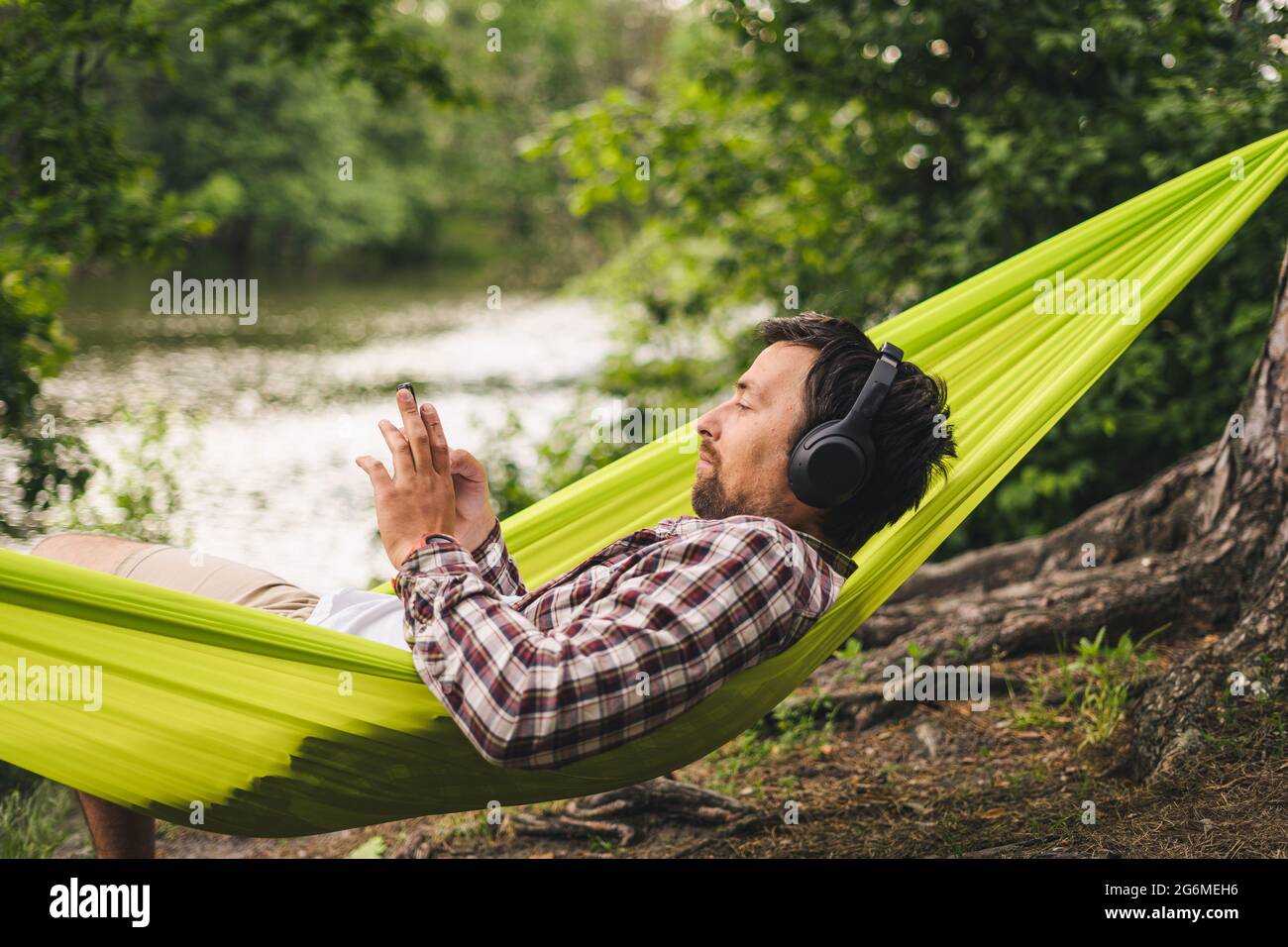 Man on bicycle trip at camping by lake is relaxing in green hammock while listening to music. Active recreation theme in nature. Hipster cyclist with Stock Photo