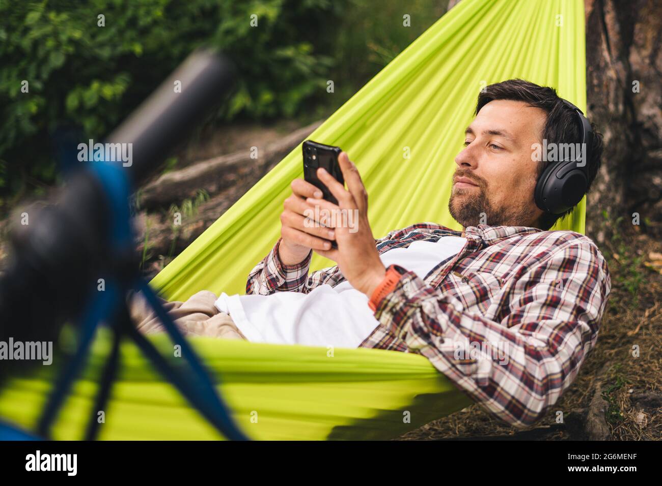 Man on bicycle trip at camping by lake is relaxing in green hammock while listening to music. Active recreation theme in nature. Hipster cyclist with Stock Photo