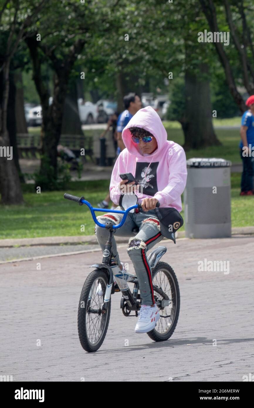 A young man in a pink hoodie reads from his phone while pedaling his bicycle. In Flushing Meadows Corona Park in Queens, New York. Stock Photo