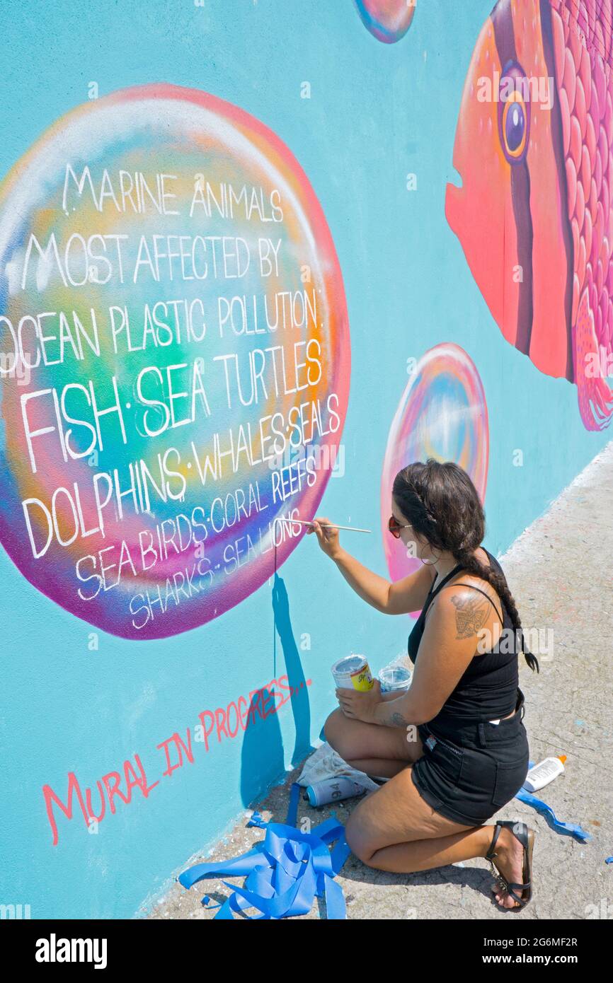 A female artist muralist writes a message about polluting the oceans with plastic on a mural on the boardwalk in Coney Island, Brooklyn, New York City. Stock Photo