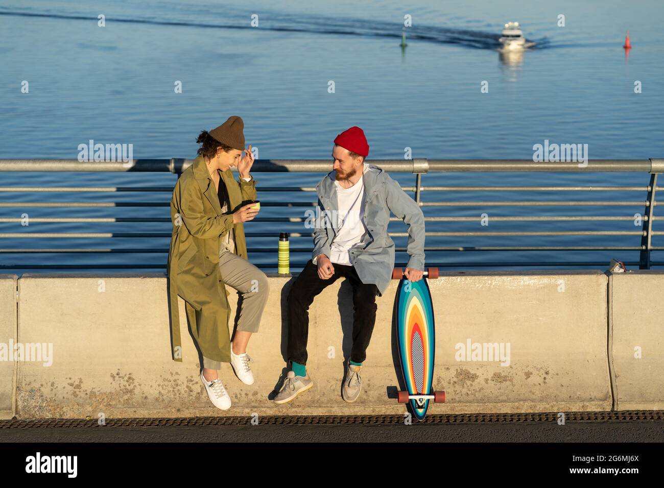 Couple of hipsters in love sit on concrete pavement of river bridge ...
