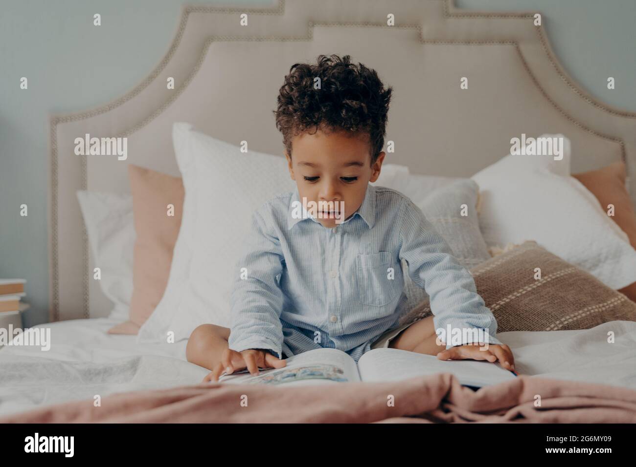 Intrigued afro american preschool child looking at colorful pictures inside of book while sitting on big bed Stock Photo