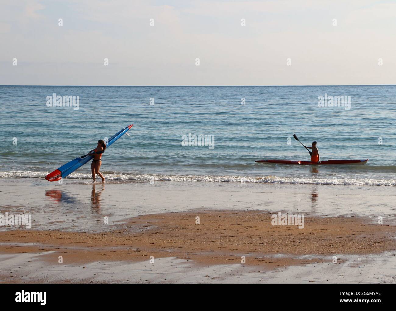 Young woman carrying a surf ski on the beach with a young man preparing to carry his out of the water Sardinero Santander Cantabria Spain Stock Photo