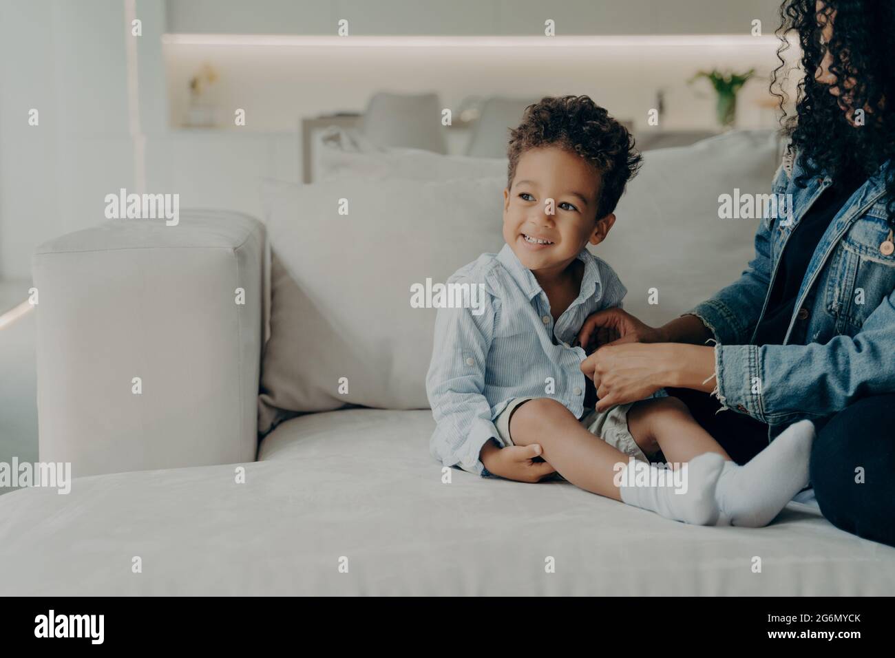 Caring afro american mom dressing up little son while sitting on sofa in living room Stock Photo