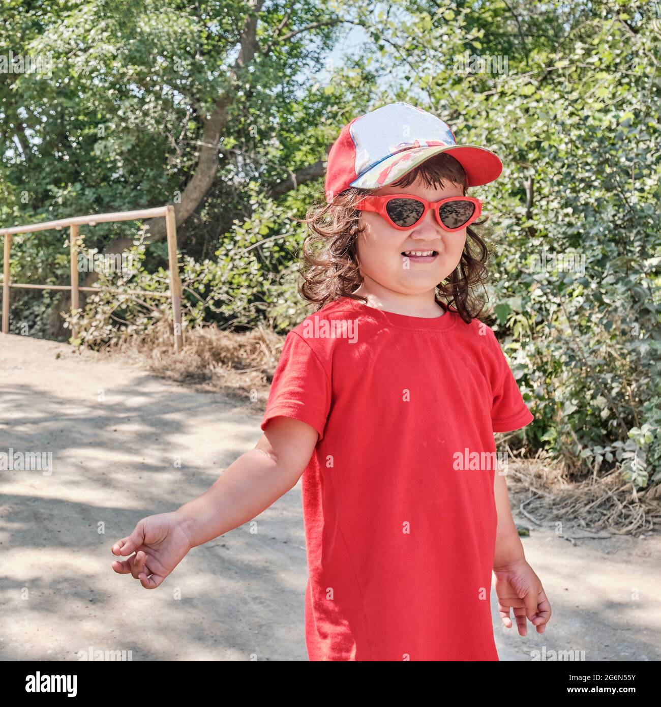 Portrait of a little Asian girl in a red dress, red sunglasses and a baseball cap walking along the road and posing. Stock Photo