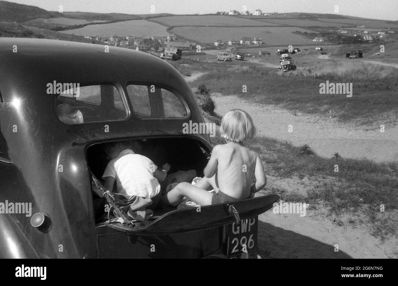 1950s, historical, two little boys in the boot or trunk of a car of the era, parked on some sand dunes, one of the boy's in the storage area, the other outside sitting on the fold down boot lid or tailgate, England, UK. Stock Photo