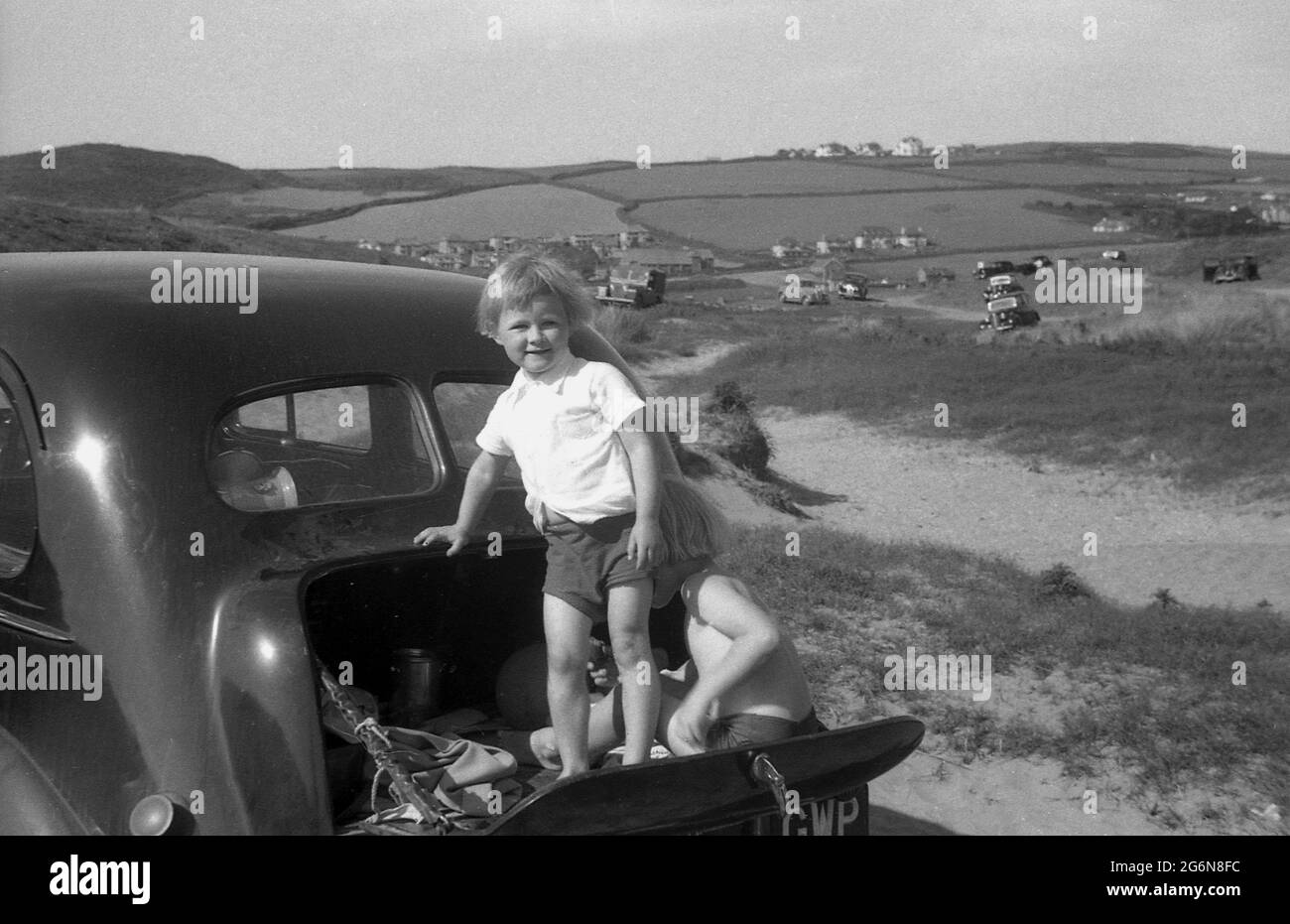 1950s, historical, two little boys in the small fold down boot or trunk of a car of the era, parked on sand dunes, one standing up on te hinged tailgate, the other sitting on it, England, UK. Other cars of the era can be seen on the dunes in the distance. Stock Photo