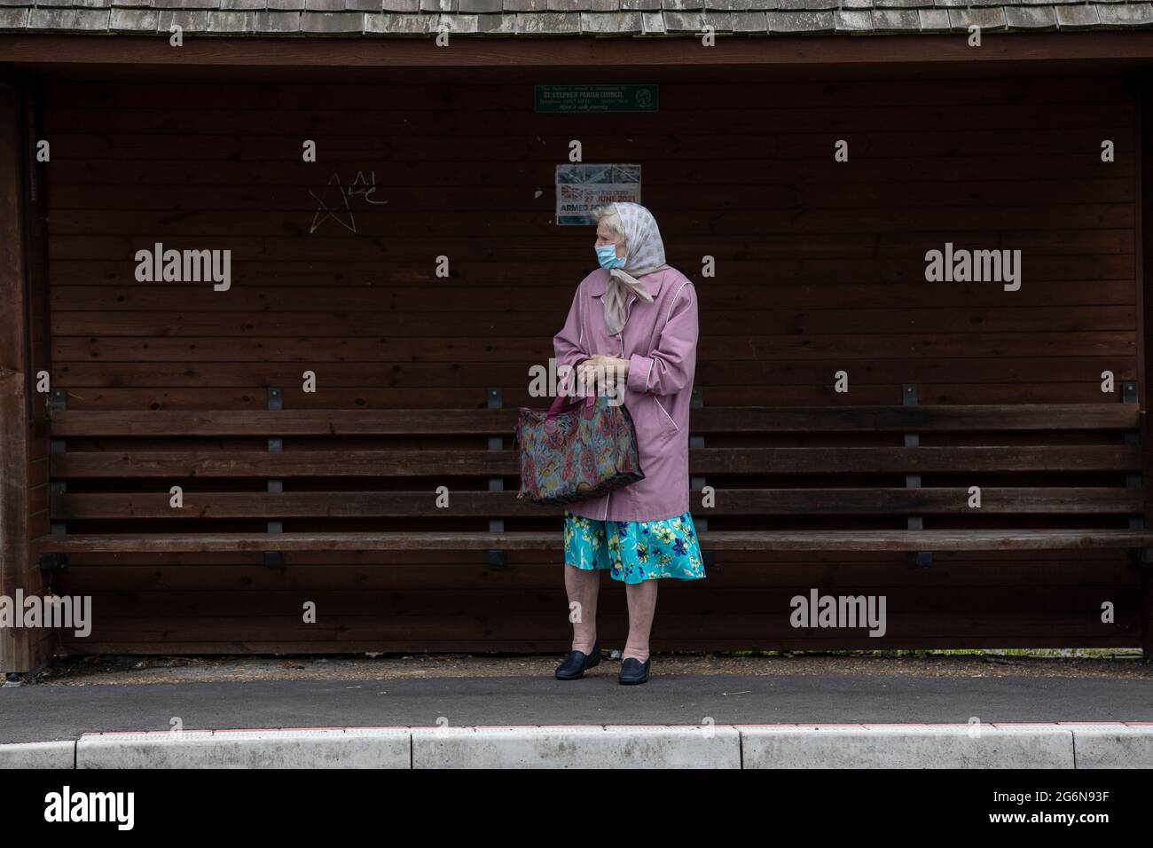 An elderly lady stood at a bus stop waiting for the next bus to arrive on a blustery day, England, United Kingdom, UK Stock Photo